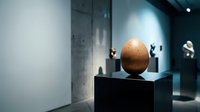Wooden egg sculpture displayed on black pedestal in modern art gallery with minimalist lighting and contemporary exhibition space for cultural presentations.