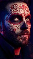 Bearded man with ornate red and gold Day of the Dead face paint featuring decorative swirls and patterns in dramatic lighting for Halloween celebrations.
