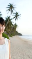 Young East Asian woman wearing white sleeveless top standing on tropical sandy beach with palm trees and ocean in background. Suitable for travel, lifestyle, and wellness content.