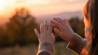 Woman raising hands toward golden sunset sky in peaceful outdoor meditation and gratitude moment during warm evening light.