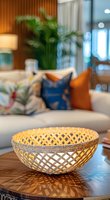 Decorative woven bamboo bowl on wooden coffee table with cozy living room setting, throw pillows and indoor plants in soft bokeh background.