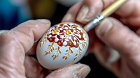 Detailed floral pattern being painted on white Easter egg using traditional wax-resist dyeing technique. Close-up view shows intricate folk art decoration process.