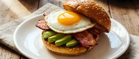 Gourmet breakfast burger with fried egg, crispy bacon and fresh avocado slices on sesame seed bun served on white plate for restaurant menu design.