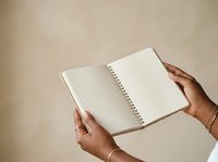 Person with medium skin tone holding open spiral-bound notebook with blank pages for writing, planning, or journaling against neutral beige background.