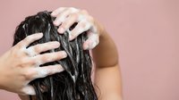 Woman washing long dark hair with shampoo foam in hands against pink background for haircare and beauty routine concepts.