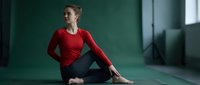 Young Caucasian woman in red top practicing yoga pose on mat in modern studio with natural light for wellness and mindfulness training.