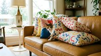 Tan leather sofa with colorful patterned pillows in modern living room with natural light from windows, wooden shelving, and floor lamp for home decor inspiration.