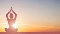 Woman practicing yoga meditation pose at sunrise with arms raised overhead creating peaceful silhouette against golden sky for wellness and mindfulness.