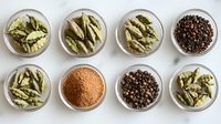 Aromatic spices in glass bowls on marble surface - green cardamom pods, black peppercorns and ground spice powder arranged in neat rows for cooking.