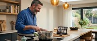 Middle-aged man with gray hair and stubble, wearing a blue shirt, cooking at a modern kitchen counter while video calling on a laptop, with pendant lights and shelving visible.