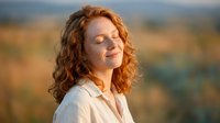 Young redhead woman with curly hair and freckles enjoying peaceful moment outdoors with eyes closed in warm sunlight for wellness and mindfulness concepts.