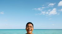 Man with dark hair and olive complexion smiling at camera in turquoise ocean water under blue sky with white clouds, summer vacation and travel concept for leisure and lifestyle use.