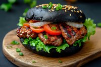 Grilled chicken burger with black charcoal bun, fresh vegetables and onion rings on wooden board. Dark moody background with green herbs garnish.