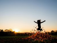 Silhouette of child jumping in pile of autumn leaves at sunset in open field with golden sky and bare trees in background.