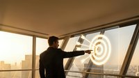 Professional businessman in suit pointing at glowing target symbol on office window overlooking city skyline at sunset for business success concepts.