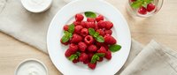 Fresh red raspberries with mint leaves arranged on white plate, overhead flat lay composition on wooden surface for food styling and culinary content.
