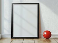 Empty black frame mockup leaning against white wall on wooden floor with red decorative ball, natural window light casting diagonal shadows.