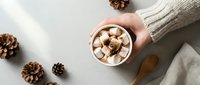 Cozy winter scene with hands holding hot chocolate topped with marshmallows, surrounded by pine cones on white wooden surface for seasonal comfort concepts.
