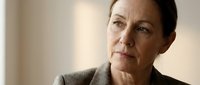 Middle-aged woman with brown hair and fair complexion, wearing gray sweater, looking thoughtfully upward in contemplative pose against neutral background.