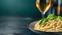 Fresh spaghetti pasta garnished with green basil leaves on dark plate, white wine glass in blurred background, moody restaurant atmosphere.