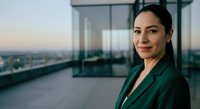 Confident female professional with dark hair and olive complexion wearing forest green blazer stands in modern high-rise office with floor-to-ceiling windows overlooking cityscape at sunset. Corporate business portrait.