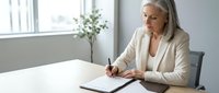 Mature businesswoman with gray hair writing in notebook at modern office desk near window, professional workplace setting for corporate presentations.