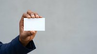 Hand holding blank white business card against neutral gray background, ideal for corporate branding and professional networking presentations.