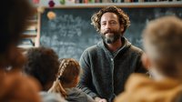 Middle-aged Caucasian male teacher with curly hair and beard smiling while teaching students in classroom with chalkboard background for education concepts.