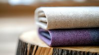 Neatly folded linen fabric in natural beige and deep purple shades stacked on rustic wooden surface, showing texture and layers in soft focus.