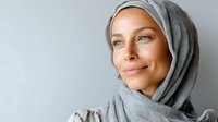 Serene young woman wearing gray hijab smiling gently against neutral background, expressing confidence and tranquility in modest fashion.