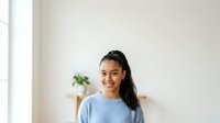 Young woman with long dark hair and medium skin tone wearing light blue casual t-shirt, smiling at camera in bright minimalist interior with potted plant and wooden shelf.