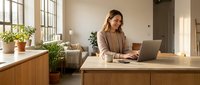 Young professional woman working on laptop in modern home office with natural light, plants and minimalist wooden desk setup for remote work productivity.