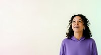 Young woman with long curly dark hair and medium brown skin tone wearing purple hoodie, looking upward thoughtfully against soft gradient background, suitable for lifestyle and motivational content.