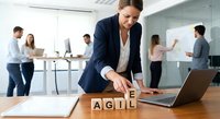 Professional businesswoman arranging wooden blocks spelling AGILE on conference table during team meeting in modern office environment.