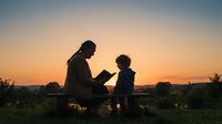 Adult male reading book to young child outdoors at sunset, silhouetted against golden sky. Suitable for education, parenting, family bonding, and literacy concepts.