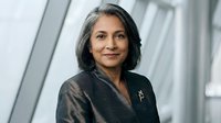 Middle-aged woman with dark hair and South Asian ethnicity wearing black blazer in modern office setting, professional portrait.