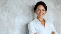 Confident Hispanic businesswoman in white shirt standing with arms crossed against textured gray wall, displaying professional leadership and success.