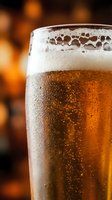 Glass of cold beer with thick foam head and condensation droplets, golden amber liquid with carbonation bubbles, blurred warm bokeh background, suitable for beverage advertising, restaurant menus, and pub promotions.