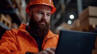 Bearded warehouse worker in orange safety hardhat and high visibility jacket using laptop computer for inventory management in storage facility.