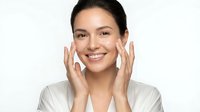Young Hispanic woman applying facial cream with gentle massage motions, smiling at camera while performing daily skincare routine in white bathrobe.