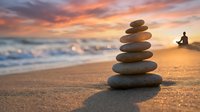 Balanced stone stack on beach at sunset with person meditating in background, zen meditation concept for wellness and mindfulness practices.