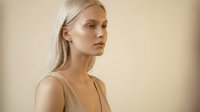 Young blonde Caucasian woman in beige tank top with natural makeup and sleek hairstyle against neutral background for beauty and fashion concepts.