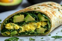 Fresh breakfast wrap filled with scrambled eggs, ripe avocado and spinach leaves in tortilla, close up view showing layers of healthy ingredients.