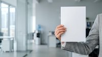 Businesswoman in gray blazer holding blank white paper sheet in modern office environment with natural light from windows.