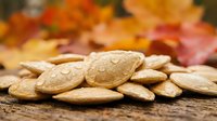 Roasted pumpkin seeds with water droplets on rustic wooden surface with colorful autumn leaves in background for seasonal cooking and healthy snacking.