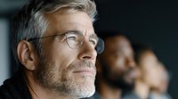 Mature businessman with glasses and gray beard smiling confidently in office environment with colleagues in background for corporate presentations.