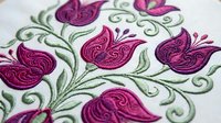 Embroidered floral pattern with purple paisley motifs and green swirls on white fabric. Traditional needlework design for textile decoration and crafts.
