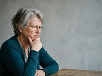 Mature woman with gray hair and glasses wearing dark blue cardigan, resting chin on hand in thoughtful pose against neutral background, suitable for senior lifestyle, healthcare, and professional concepts.
