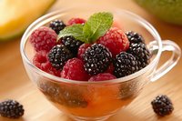 Fresh raspberries and blackberries in glass bowl with mint garnish on wooden surface, close up view of mixed berries dessert in natural lighting.