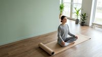 Young Asian woman in gray activewear practicing meditation in modern minimalist room with yoga mat, plants and natural light for wellness lifestyle.
